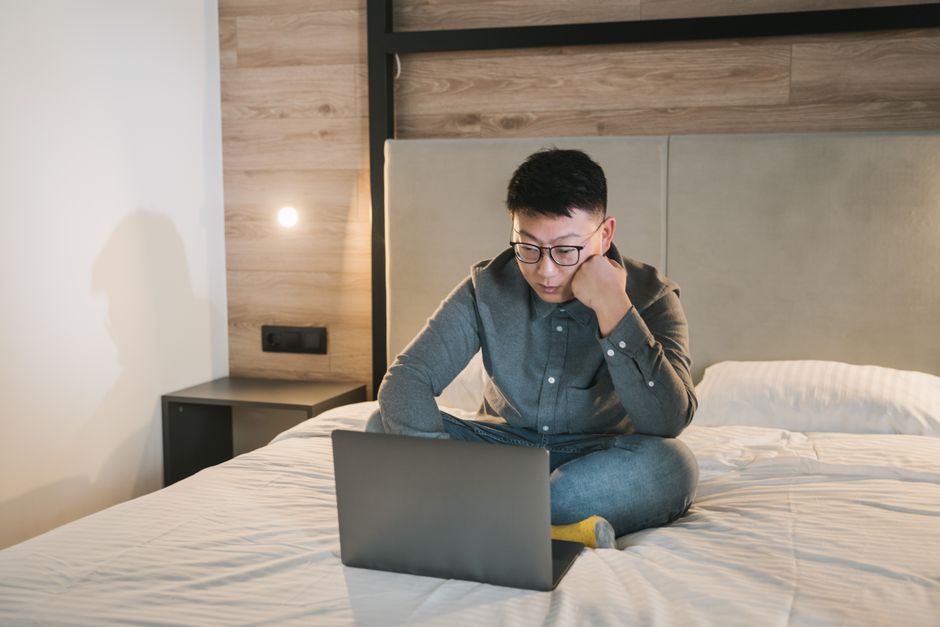Asian man with glasses sitting on a bed, focused on his laptop, working remotely.
