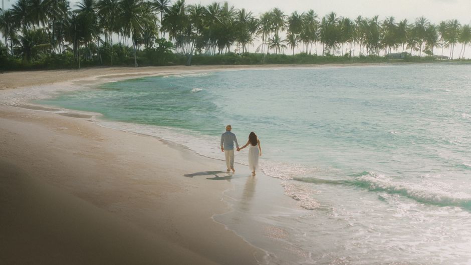 Couple walking hand in hand on a sunny Cancún beach, vibrant palm trees and ocean.