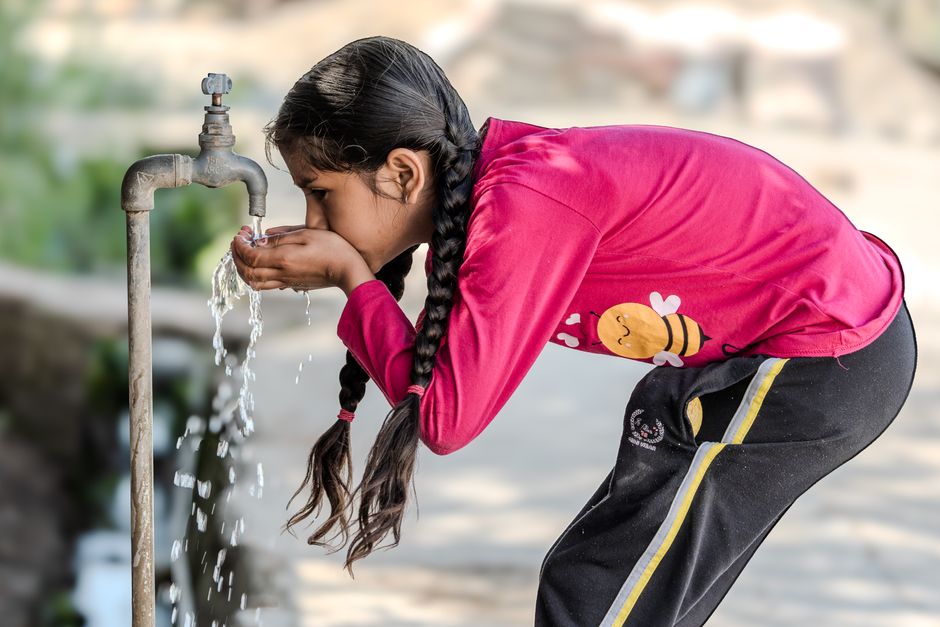 A young girl drinks water from an outdoor tap on a sunny day, emphasizing the simplicity of rural life.