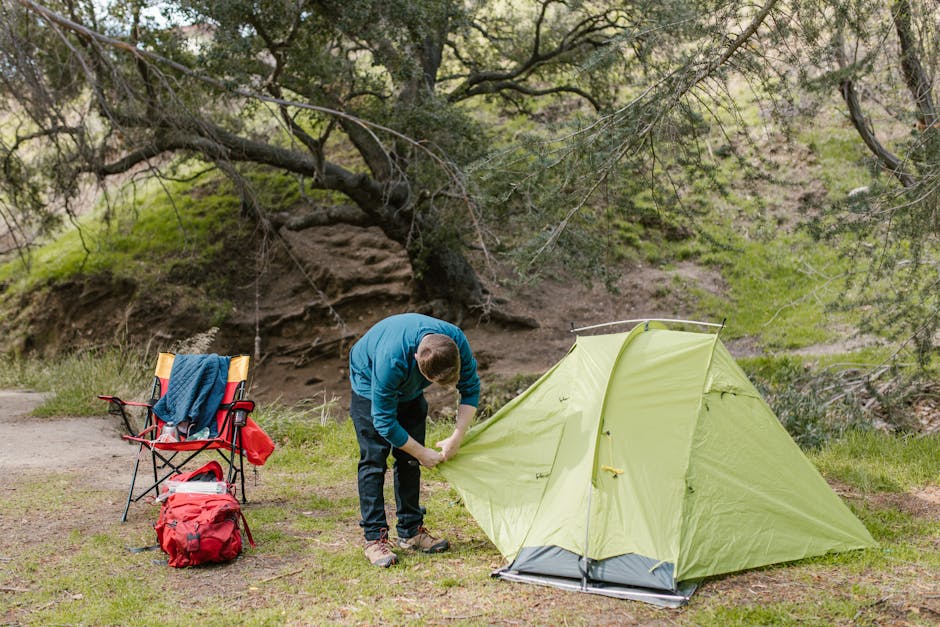 A man in blue sets up a green tent in a peaceful outdoor setting with a chair and backpack.