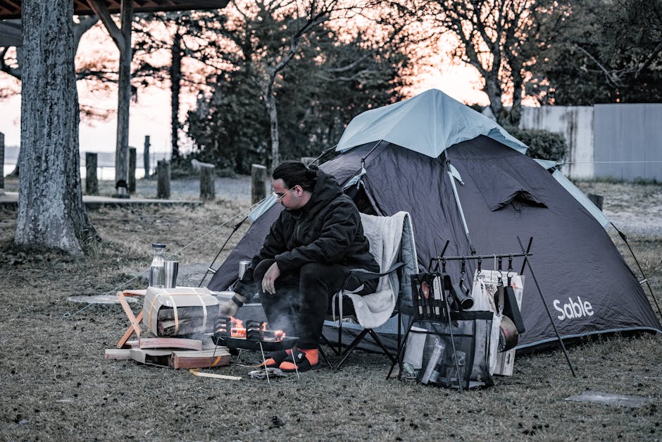 A man enjoys a campfire beside his tent amid a tranquil forest during sunset.