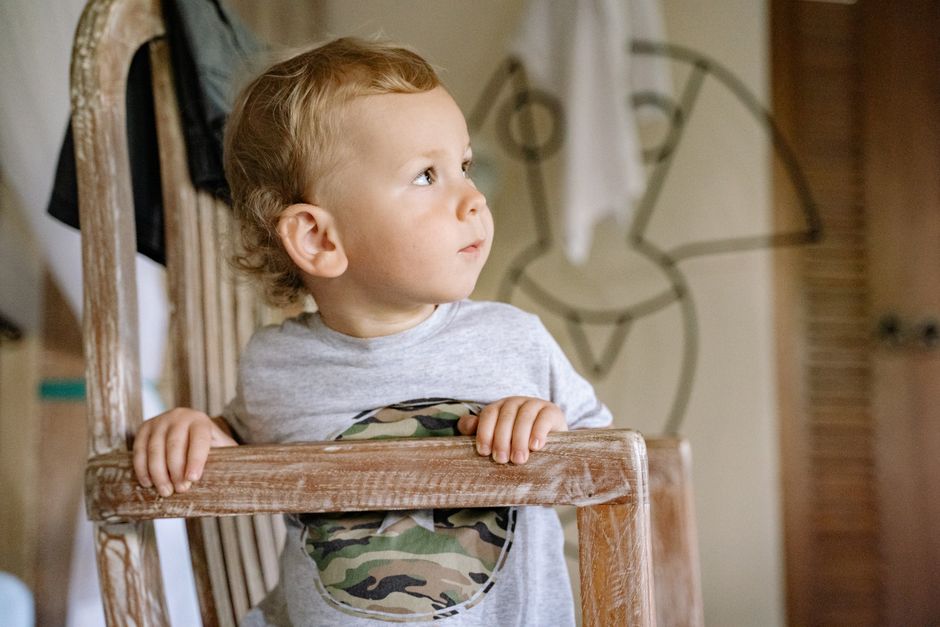 A toddler sits on a wooden chair indoors, gazing curiously to the side, in a calm and natural setting.