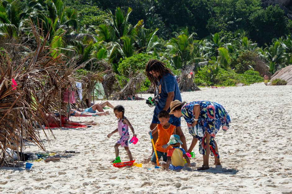A family enjoys a day at a tropical beach, playing in the sand surrounded by palm trees under a bright summer sun.