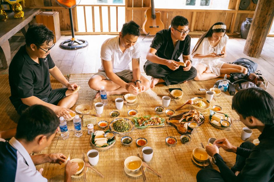 A group of six adults enjoys a communal meal indoors, seated on a mat.