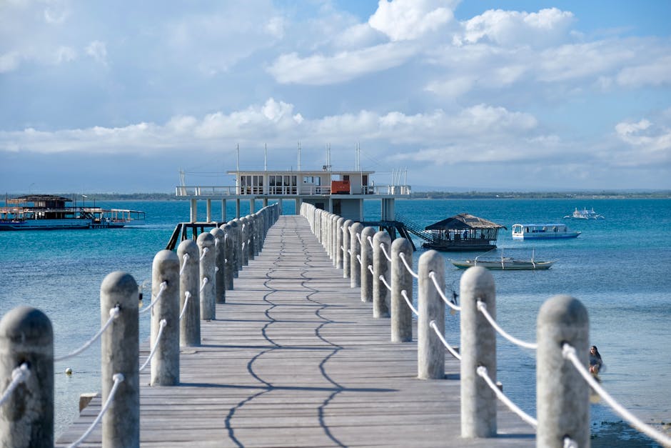 A long wooden pier stretches into the blue sea at a tropical resort, featuring boats and clear skies.