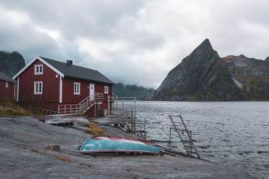 Quaint red cabin by Norwegian fjord with dramatic mountain backdrop under cloudy skies.