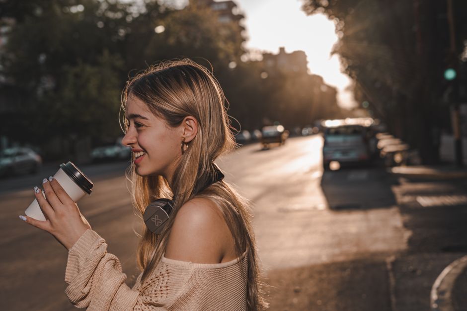 Smiling young woman with headphones and coffee on city street at sunset.
