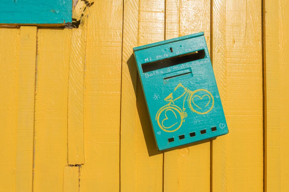 Teal mailbox with bicycle design against a bright yellow wooden wall in sunlight.